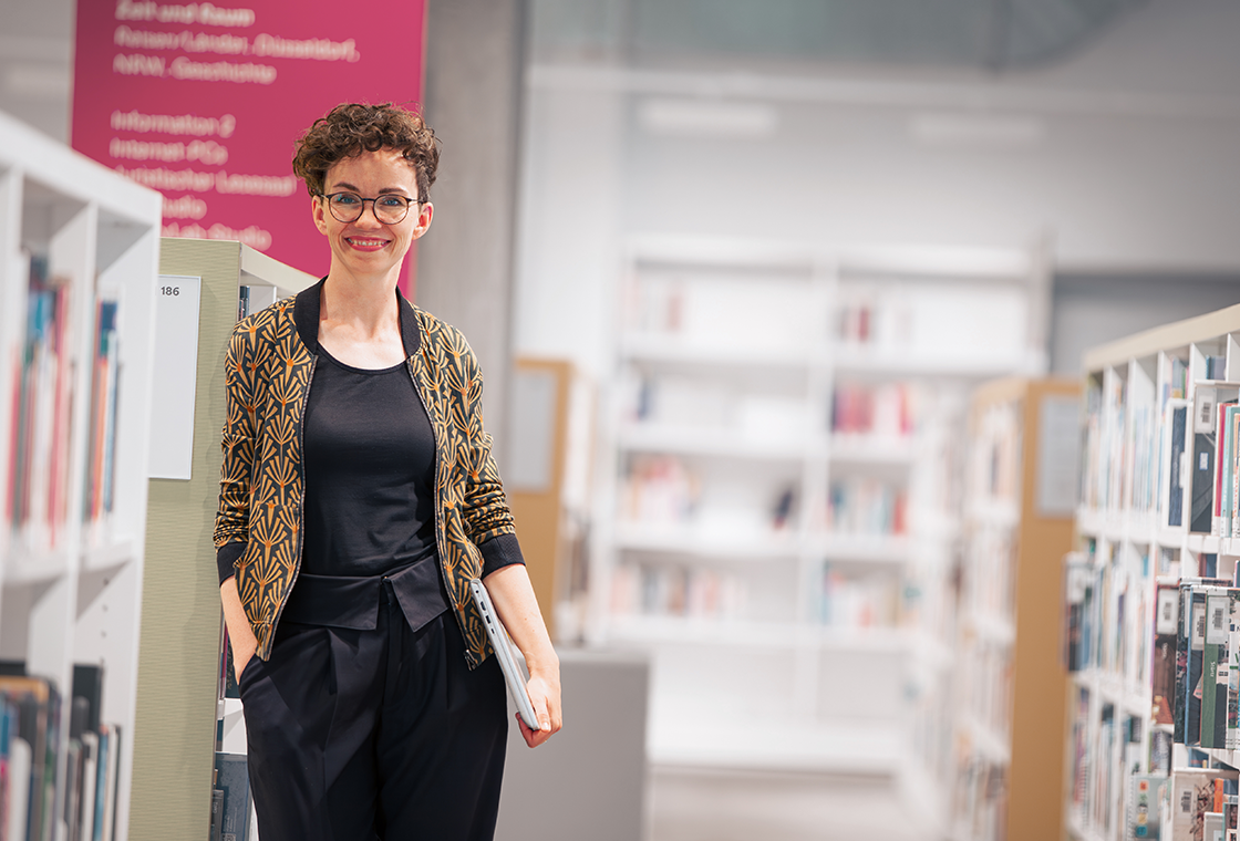 Verena Smit lehnt sich in einer Bibliothek an ein Bücherregal und schaut mit Laptop unter ihrem Arm direkt in die Kamera.